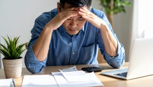 机に広げた請求書を前に頭を抱える男性、請求書払いの負担や支払い期日に悩む様子
