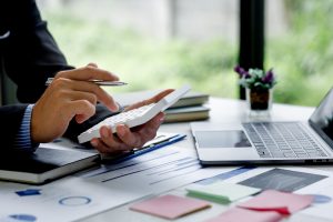 デスクに書類やグラフを広げ、電卓とペンを使って手作業で数字の確認を行っている様子。請求書処理の自動化によって効率化できる、従来のアナログな経理業務のイメージ。