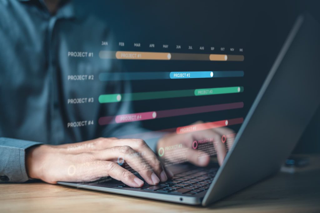 ノートパソコンの画面上に浮かぶプロジェクト進行表のホログラムを操作する手元。助成金の申請期限や対象となる事業計画のスケジュールを管理している様子