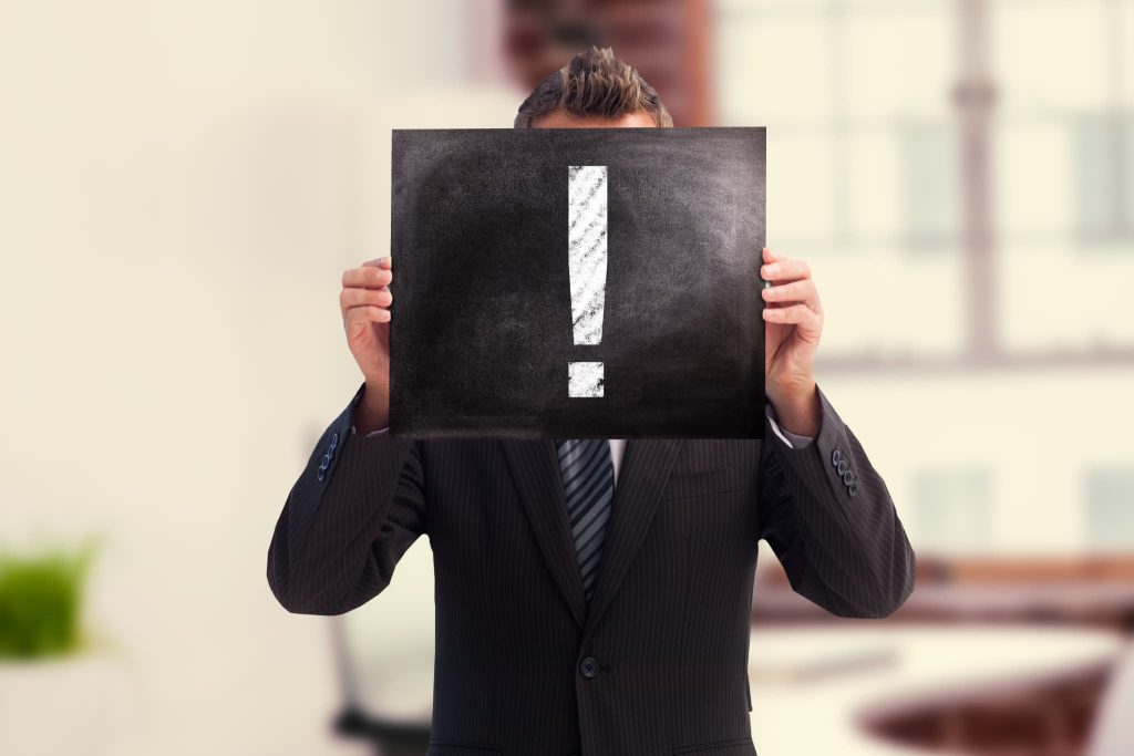 「！」マークが描かれたボードを顔の前に掲げるビジネスマン。当座貸越とは何かを理解する上で押さえておくべき注意点やリスクを示唆するイメージ