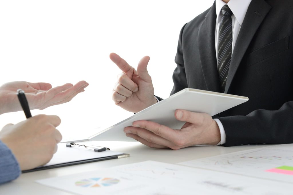 タブレット端末を持ったスーツ姿の担当者が、対面する相手に身振りを交えて説明している。銀行融資の面談やビジネスローンなど、対話を通じた運転資金の調達プロセスや交渉の場面