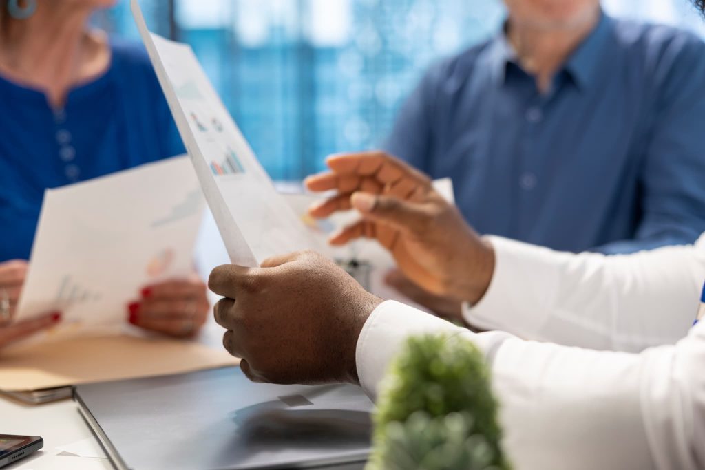 グラフが描かれた資料を手に持ち複数の関係者がファクタリングのメリットデメリットや契約内容について議論しているビジネスミーティングの様子