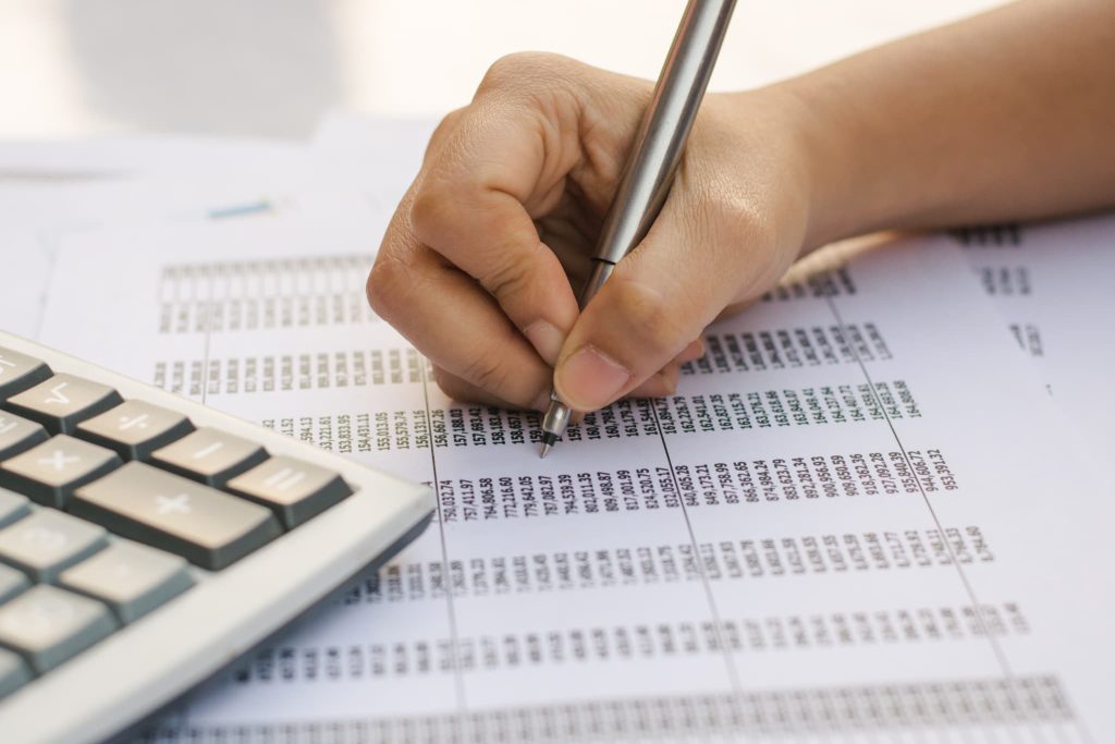 数字が並んだ帳簿にペンでチェックを入れ電卓を使っている手元。売掛金とは日々の取引を記録し入金消込を行う経理実務において重要な管理項目