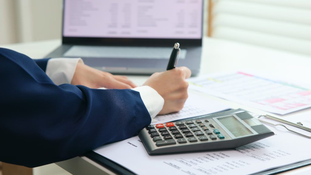 電卓を脇に置き書類に数字を記入している経理担当者の手元。当座貸越とは資金不足の際に自動で融資を受けられるため資金繰り管理に役立つ仕組み