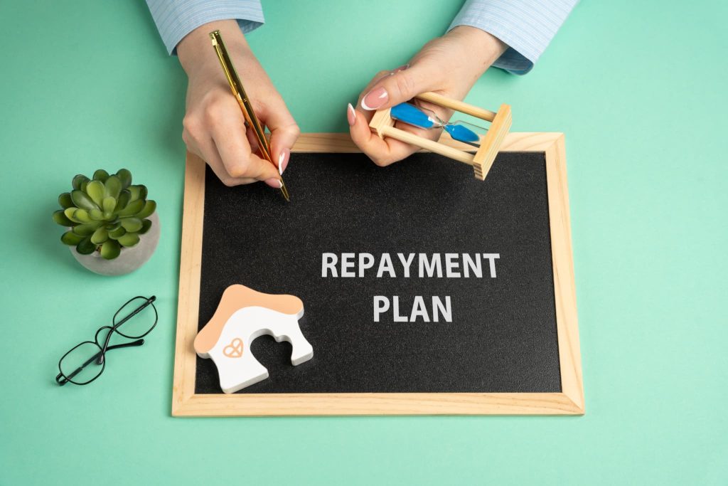 ファクタリングの返済計画を立てるために砂時計や家のオブジェを並べREPAYMENT PLANの文字を書く様子