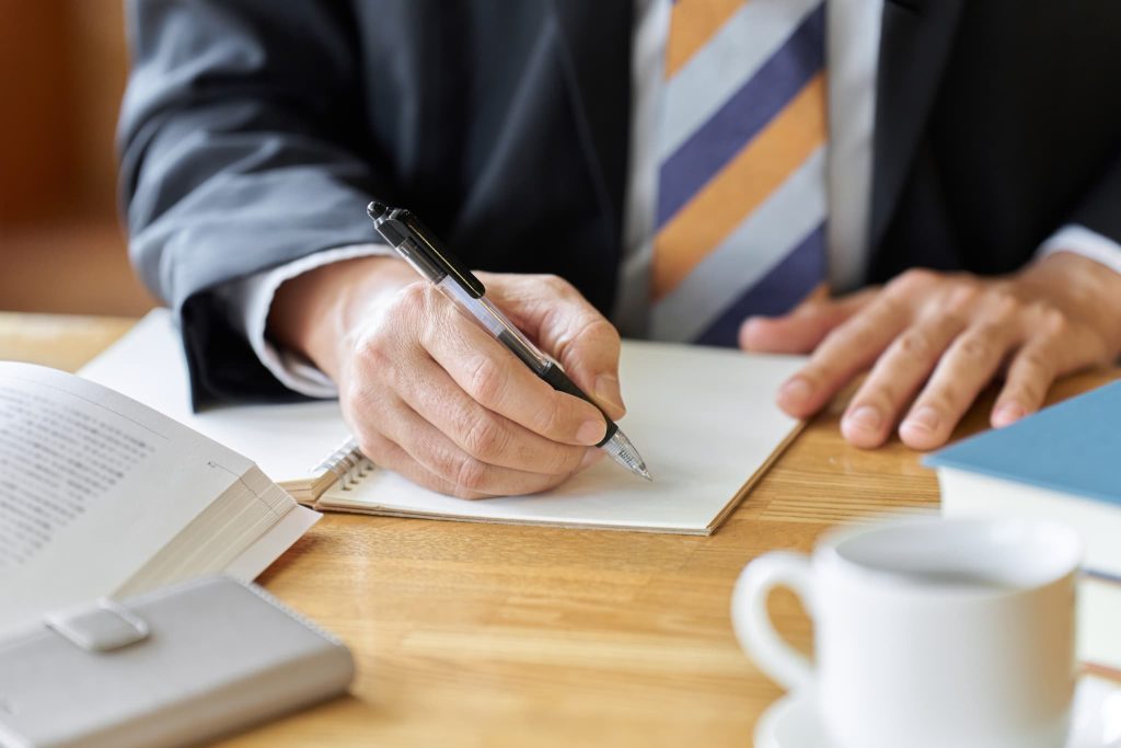 コーヒーを傍らに置きノートにペンを走らせている男性の手元。ビジネスローンの審査申し込みに必要な書類を準備したり、即日融資に向けて計画を練っている様子