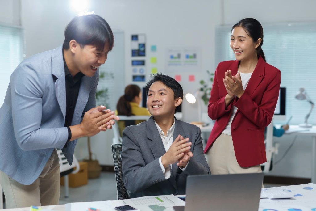 パソコン画面を見ながら笑顔で拍手し合う3人のビジネスチーム。事業資金の融資審査が承認され、プロジェクト始動に向けた資金確保の成功を喜び合っている活気あるオフィスの様子