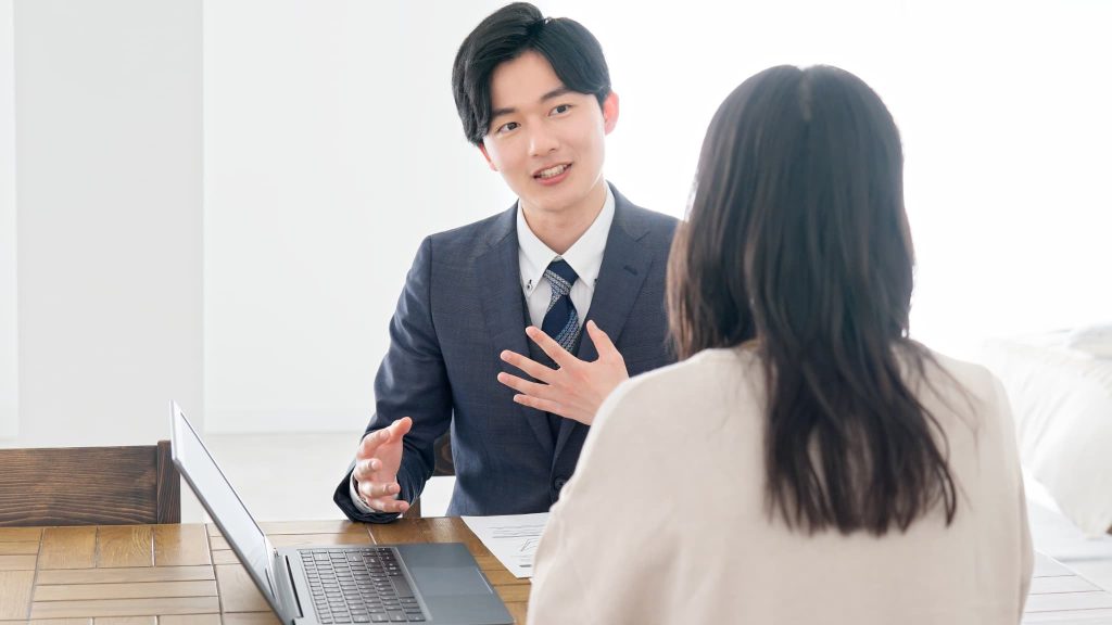 ノートパソコンを開き、対面する相手に熱心に説明を行うスーツ姿の男性。銀行窓口での事業資金の融資相談や、事業計画書に基づいた資金調達の交渉を行っている真剣なビジネスシーン