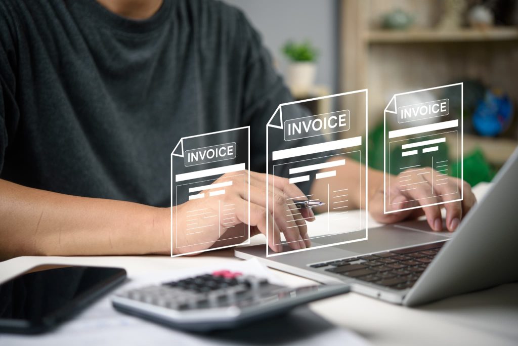 ノートパソコンと電卓を使い、画面上に浮かぶデジタルのINVOICEアイコンを確認しながら経費精算業務を行う手元。立て替え払いとは何かに関連する請求書処理や経理のイメージ