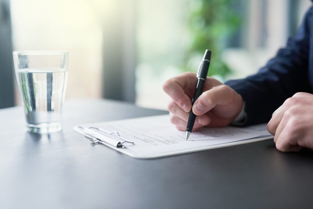 デスクの上で契約書類にペンを走らせる男性の手元。短期借入金や長期借入金の融資実行に向けて契約手続きを進めているビジネスシーン