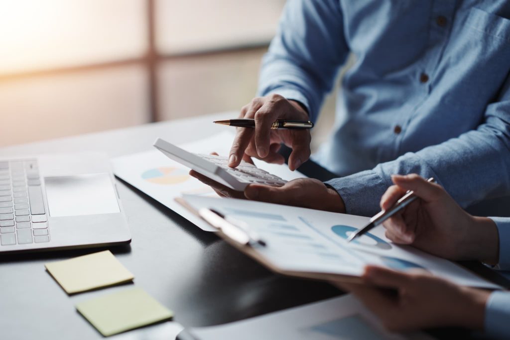 電卓を操作して数値を計算する人物と、資料のグラフをペンで指す人物。綿密な資金繰り計画やコスト管理を通じて、着実にキャッシュフロー改善を進める業務風景