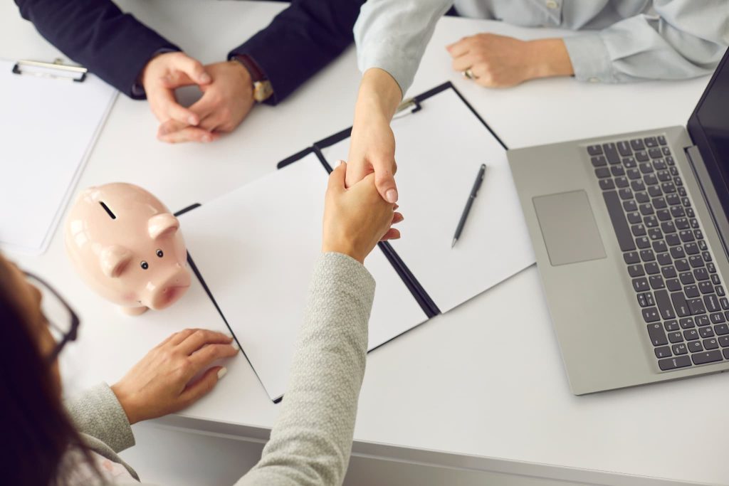 契約書の上で握手を交わす手元と、テーブルに置かれたピンクの豚の貯金箱。事業資金の融資契約が成立し、資金調達に成功した瞬間や、金融機関とのパートナーシップを象徴している