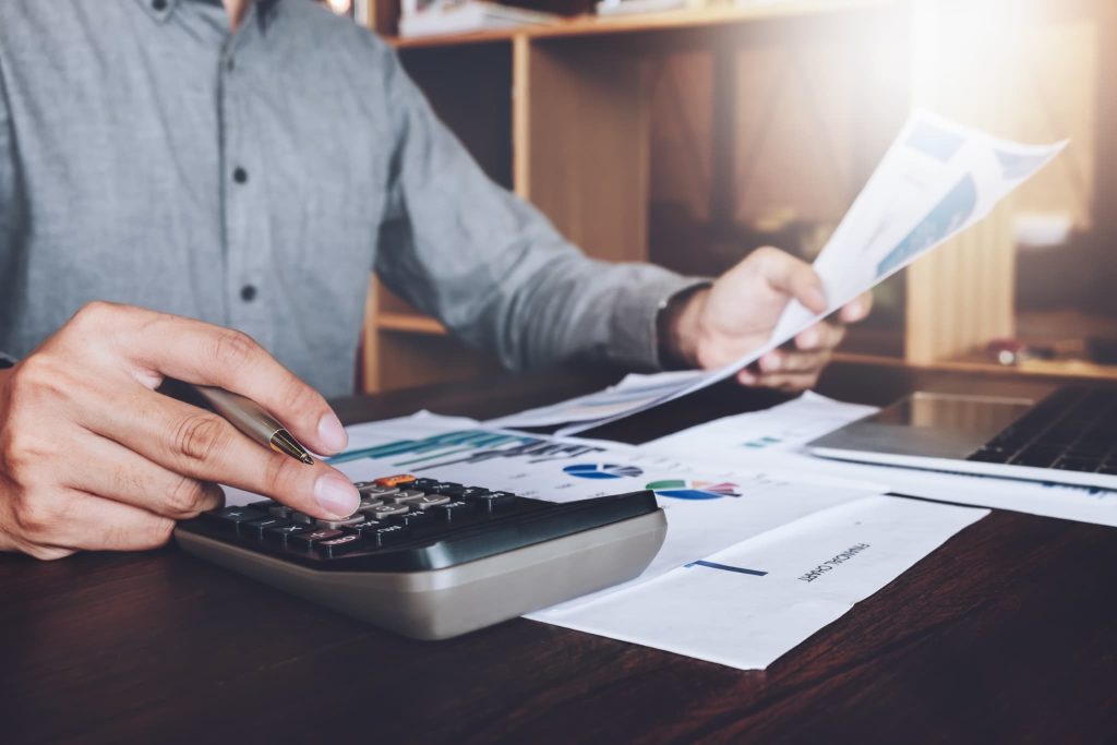 グラフ入りの資料を見ながら電卓で計算を行う男性の手元。飲食店の事業計画書作成や収支シミュレーションなど、資金調達に向けた数値計画の策定風景