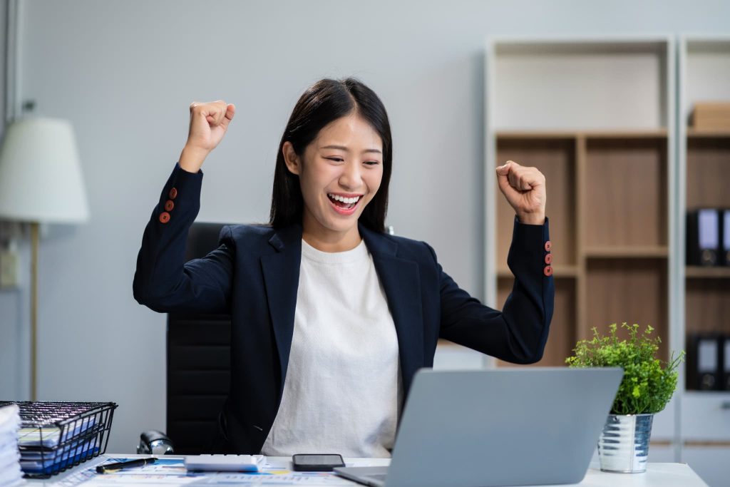 パソコンの前で両手でガッツポーズをして喜ぶ女性。ビジネスローンの審査に通過し、即日融資などのメリットを受けて資金調達に成功した様子