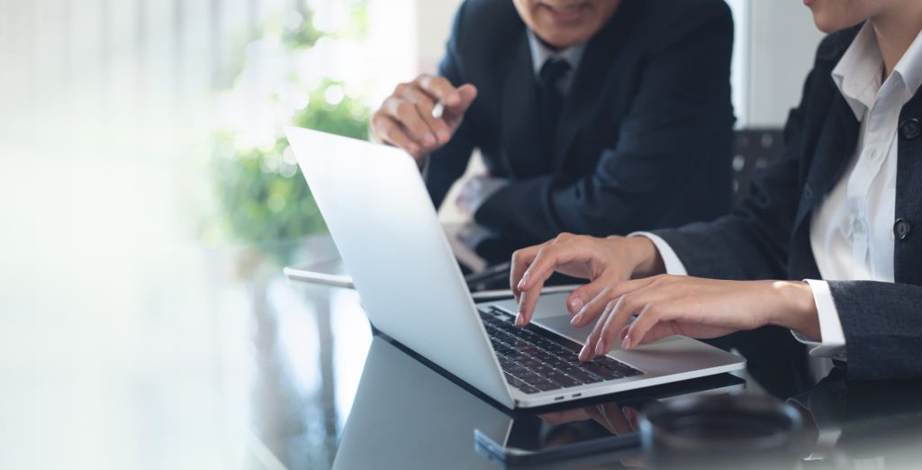 ノートパソコンの画面を見ながら話し合うスーツ姿の男女。プロパー融資の審査対策として事業計画書を作成したり銀行員と相談している様子