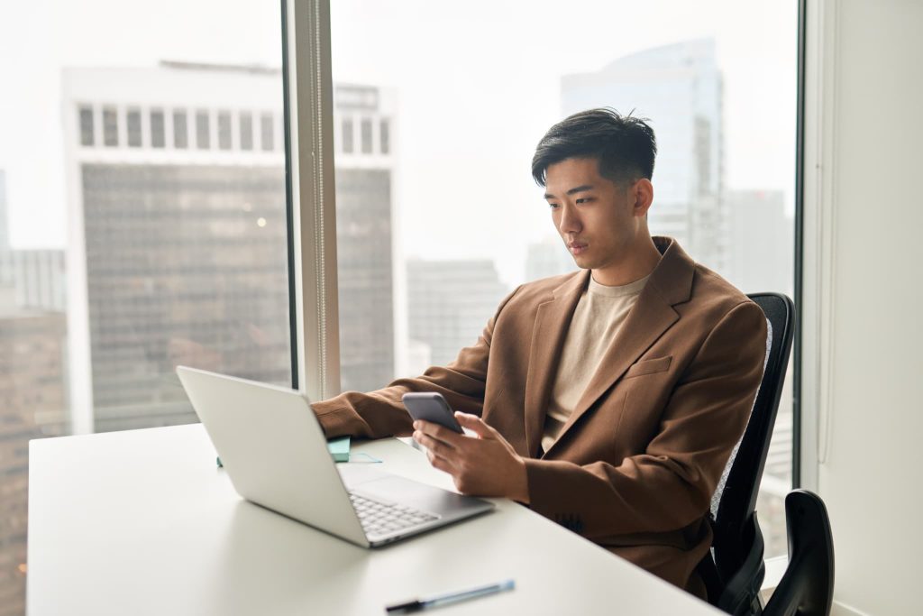 オフィスの窓際でノートパソコンを開きながらスマートフォンを操作してファクタリングの申し込みや入金確認を行っている男性