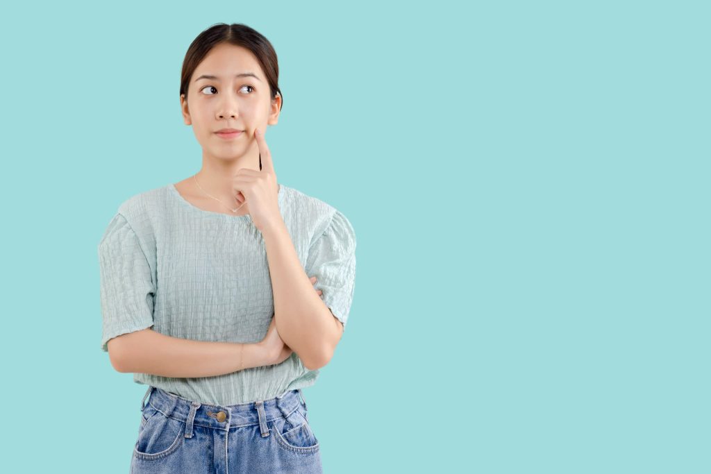 指を頬に当てて上を見上げ疑問を抱いている女性。請求書払いとはどのような仕組みか学んだり導入を検討している様子