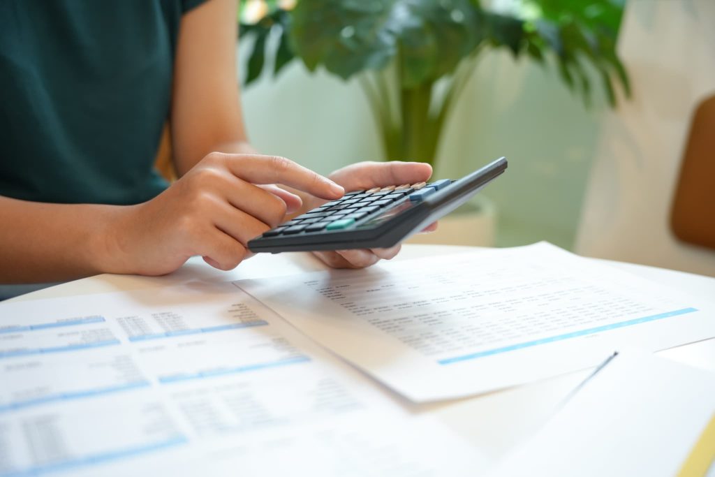 書類が広げられたデスク上で黒い電卓を使い、掛取引における支払いサイトの期間確認や経理処理を進めている女性の手元のクローズアップ