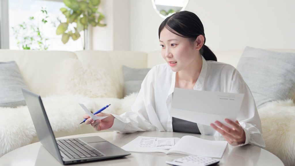 請求書を手に持ちパソコン画面を確認する女性。ファクタリングの手数料相場を調査し、資金調達コストを試算している経理業務