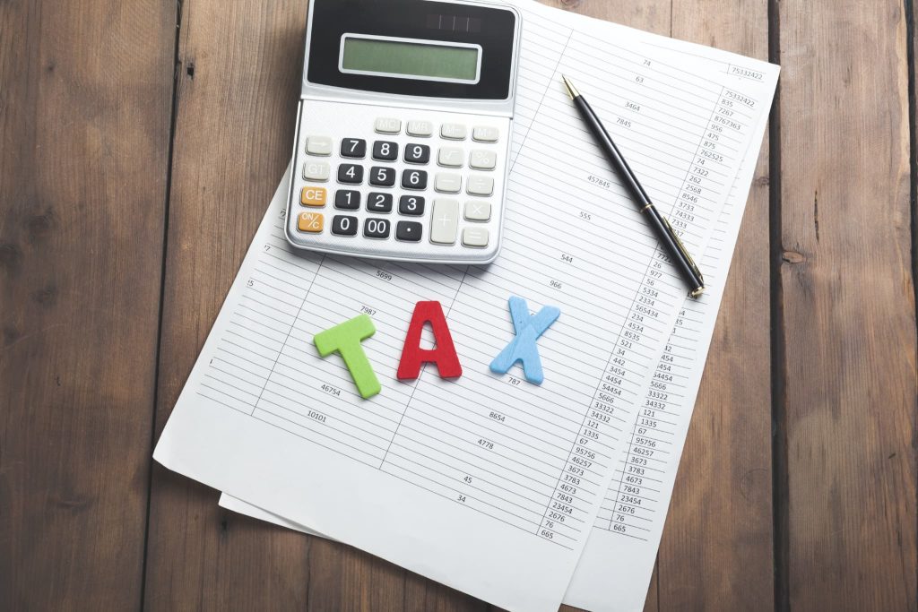 数字が羅列された書類の上に置かれたTAXの文字ブロックと電卓。助成金は原則として課税対象となるため税務処理や確定申告が必要であることを示すイメージ