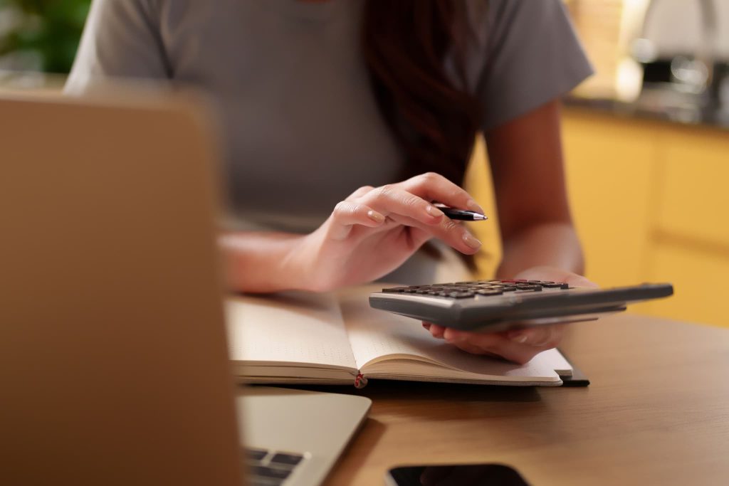 手帳とパソコンを広げ、電卓を手に持つ女性。帳簿付けや経費の計算を行うことで、事業に必要な運転資金を管理し、健全な経営状態を維持しようとする実務風景