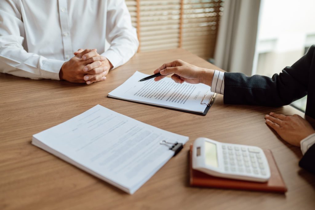 デスク上の契約書に向けてペンを差し出しファクタリング契約の手続きや対面での審査説明を行っているビジネスマンの手元