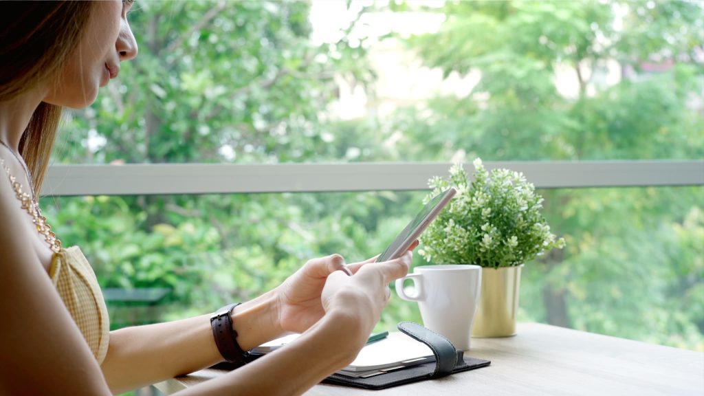 窓辺で手帳を広げスマートフォンを操作する女性。短期借入金や長期借入金の口座残高を確認したり返済シミュレーションを行っている様子