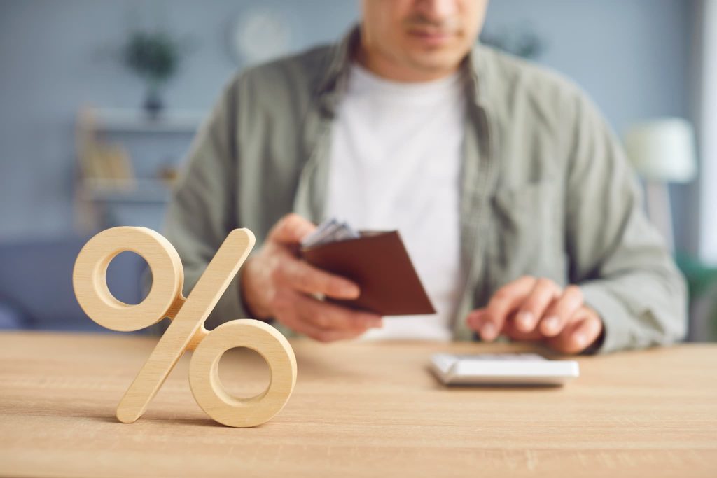 木製のパーセント記号のオブジェと背景で電卓を持つ男性。プロパー融資の金利条件を確認したり返済利息の計算をしているイメージ