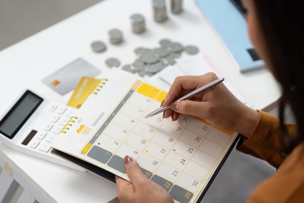 小銭やクレジットカードが置かれたデスクでカレンダーの日付を確認し、支払いサイトを考慮した入出金スケジュールの管理を行っている様子