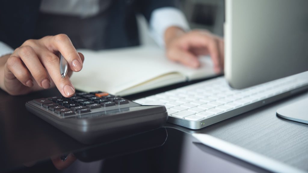 オフィスのデスクでキーボードを前に置き電卓を叩く手元。助成金の申請に向けた正確な数値計算や資金計画を練っているビジネスシーン