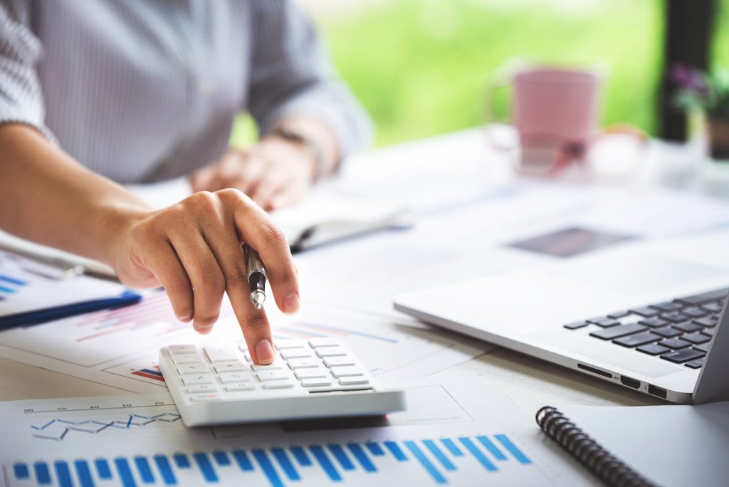 資料やグラフを広げたデスクで電卓を操作している女性。助成金の申請に必要な経費を算出したり受給額の試算を行っている経理業務