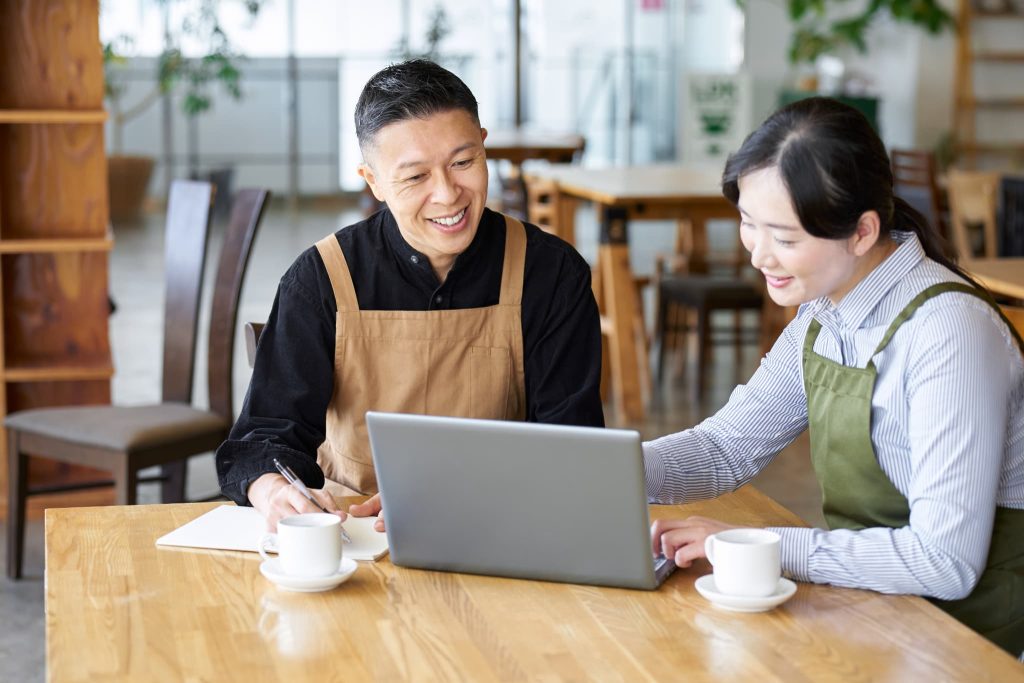 エプロン姿の飲食店オーナーとスタッフが、パソコンを見ながら笑顔で相談している。店舗運営の状況を確認し、将来の改装や設備投資に必要な運転資金について話し合っている様子