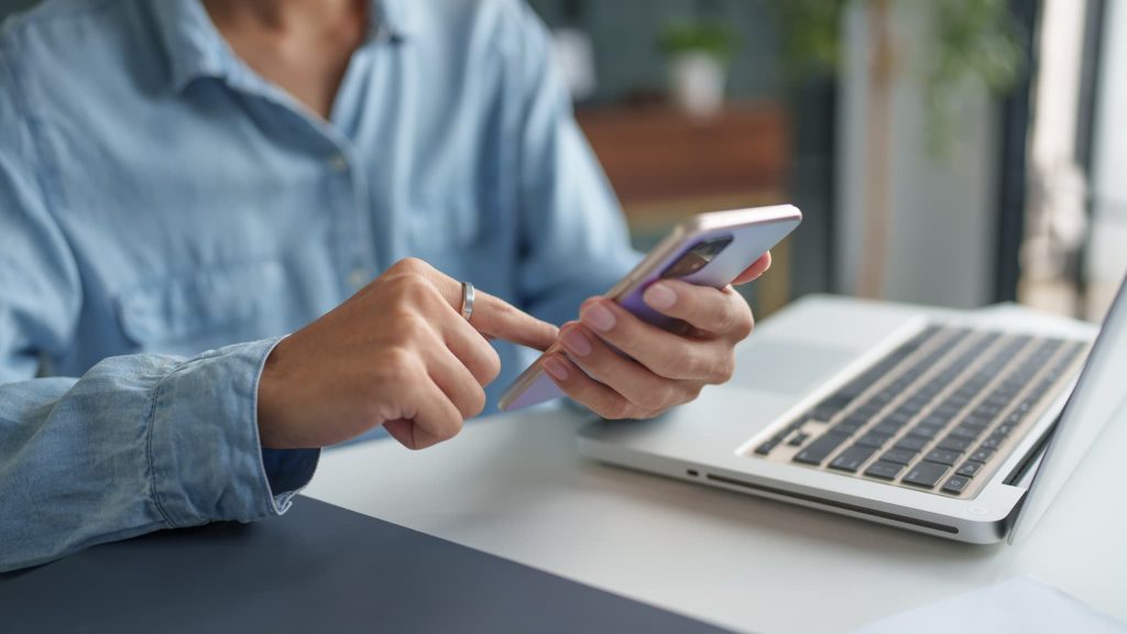 ノートパソコンの前でスマートフォンを操作している手元。売掛金とは期日までに入金されたか銀行口座を確認したり取引先と連絡を取る必要がある債権