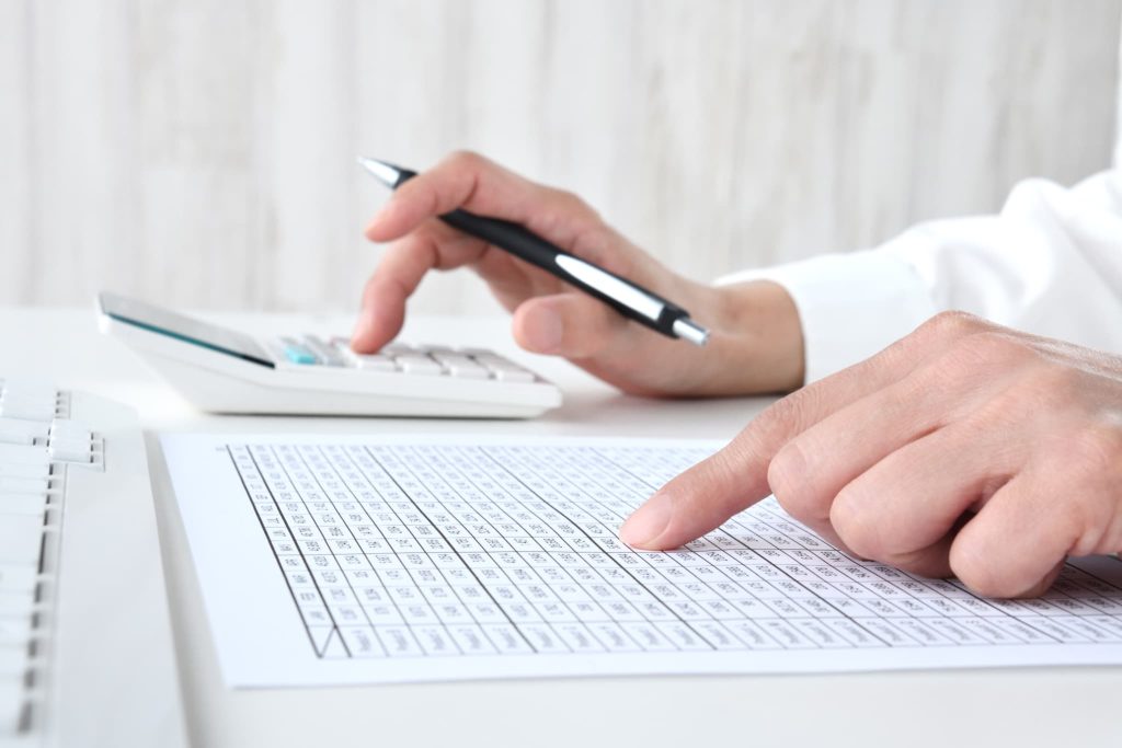 数値が羅列されたリストを指で確認しながら電卓を使う様子。綿密な資金繰り表の作成やデータ分析を行い、ショートを防ぐための適切な運転資金管理を実施している