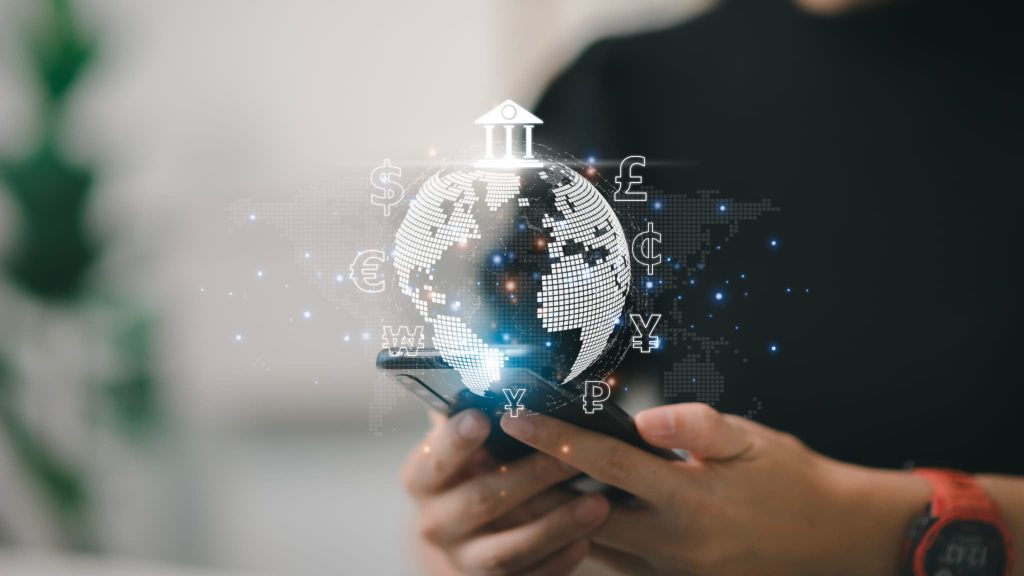 スマートフォンを持つ手の上に地球儀や円、ドルなどの通貨記号、金融機関のアイコンがホログラムで浮かんでいる様子。ノンバンクを含むフィンテックやグローバルな金融サービス