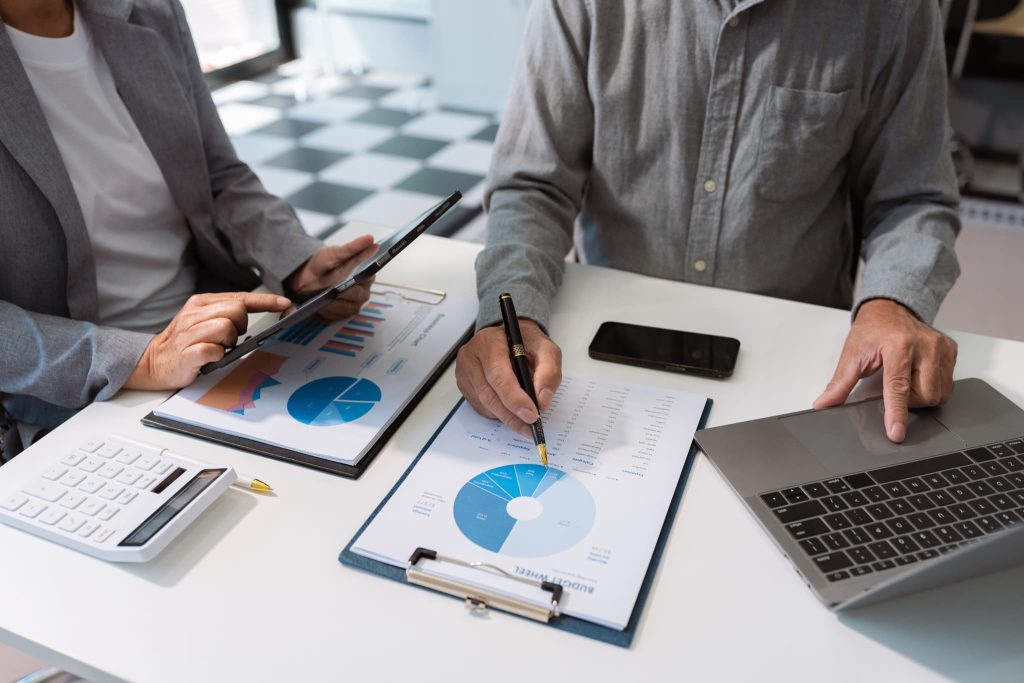 デスクでタブレットとノートパソコンを使い円グラフの資料を分析する二人。新創業融資制度の審査で重要となる市場調査や売上予測を立てるイメージ