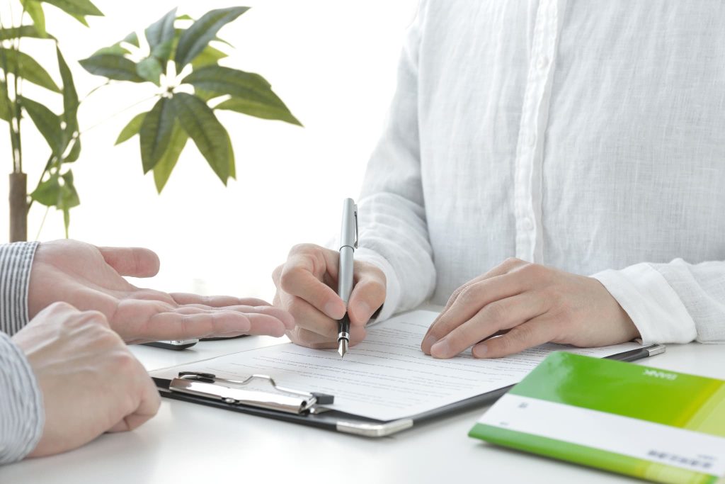 観葉植物のある明るいオフィスで書類にサインをする手元。新創業融資制度の契約手続きや、創業計画書の最終確認を行うビジネスの場面