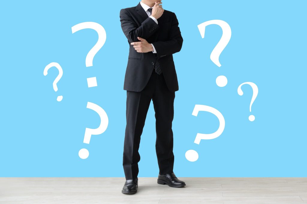 背景に大きなクエスチョンマークが並ぶ中で考え込むスーツ姿の男性。開業資金がいくら必要なのか、何から準備すべきか悩む起業家の様子