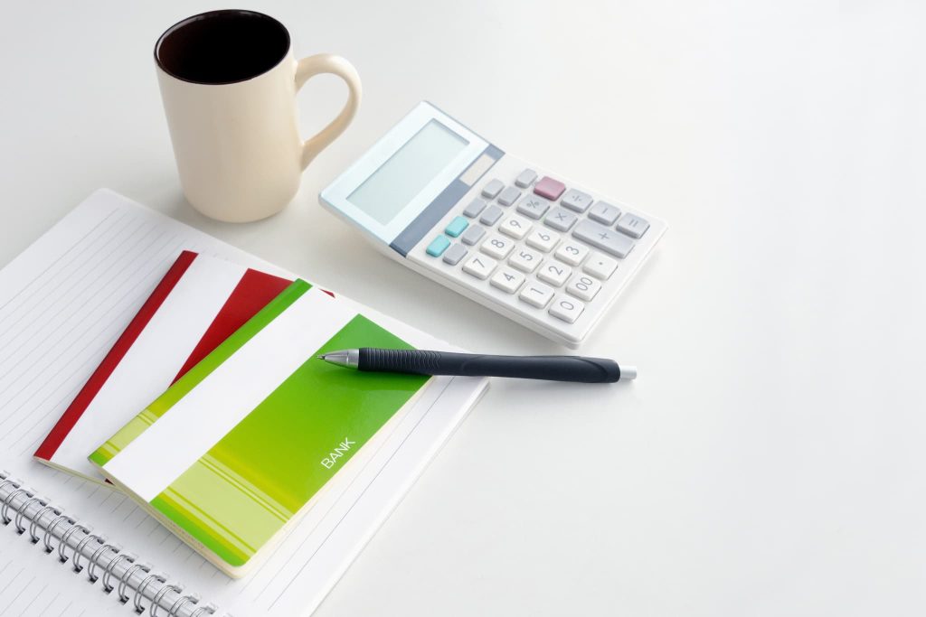 白いデスクに置かれた通帳と電卓、ボールペンとコーヒー。ファクタリングの必要書類を準備する事務作業のイメージ
