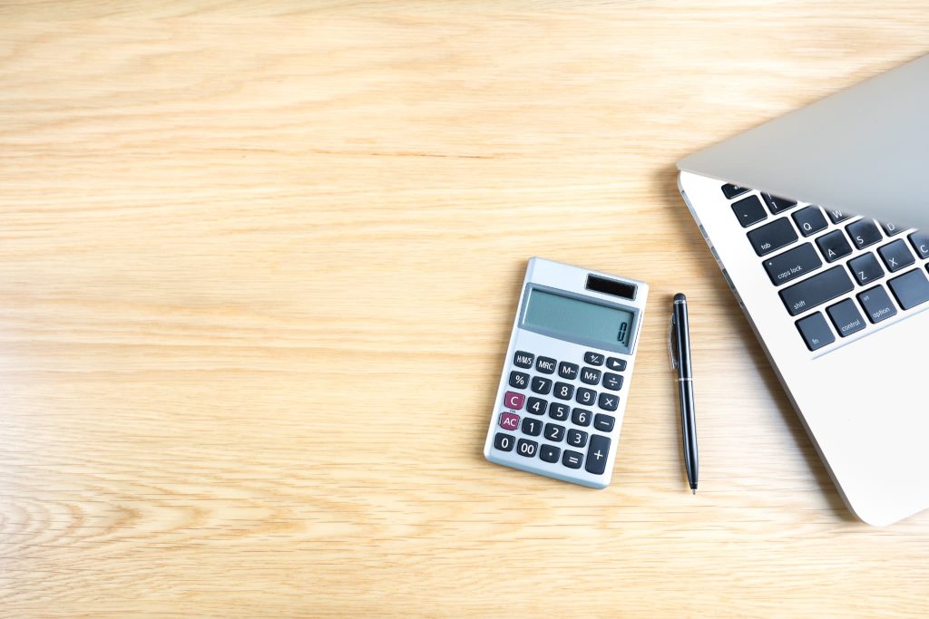 木製デスクに配置されたノートパソコン、電卓、ボールペンの俯瞰。新創業融資制度の収支計画作成に必要なツールが揃った、起業準備のデスクワーク