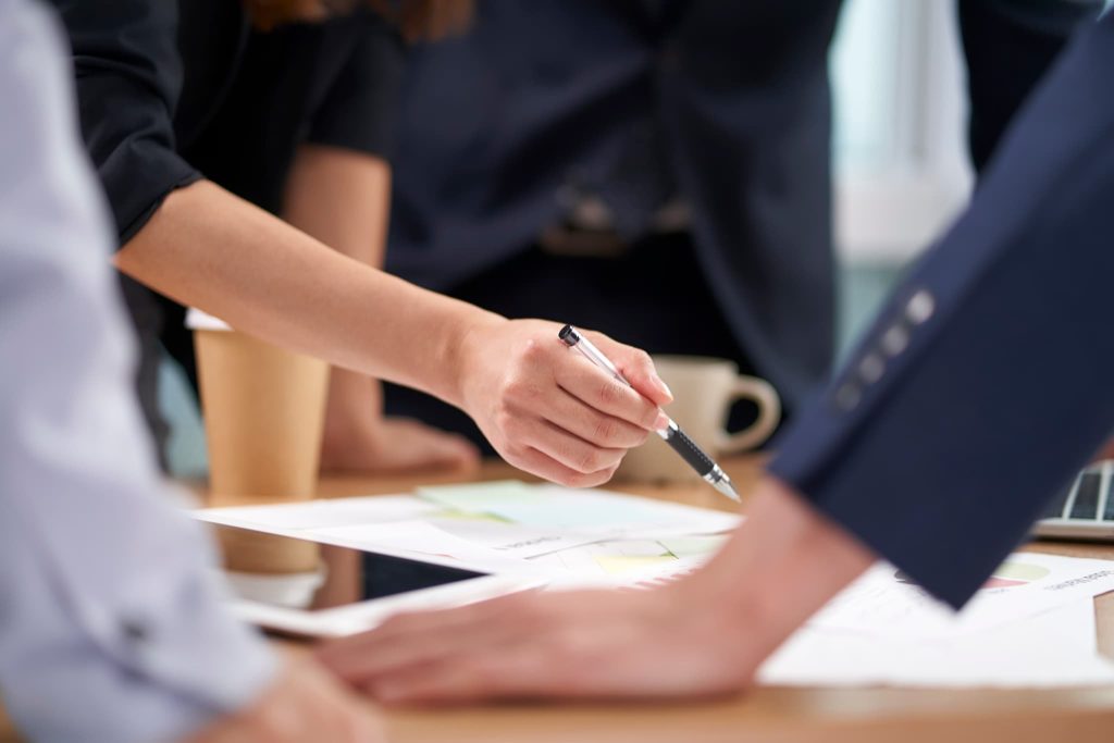 会議室で複数のビジネスパーソンが資料を囲みペンで指し示す手元。新創業融資制度を活用した事業拡大や組織的な事業計画立案のチームワーク