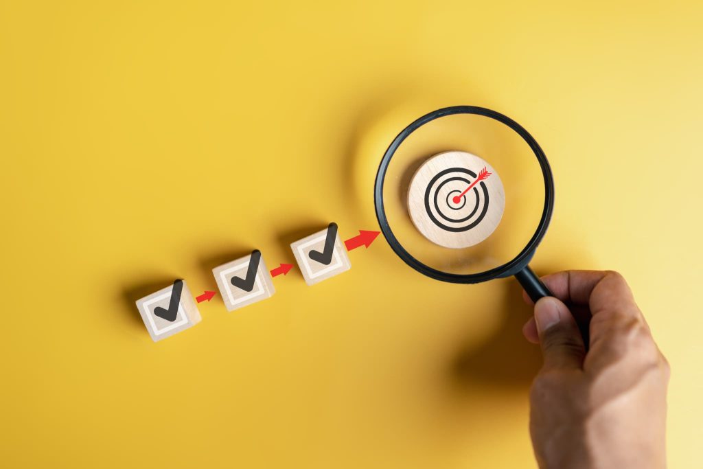 チェックボックスの先に的があるイラストを虫眼鏡で覗く手。経費削減のターゲットを明確に定め、効率的に目標を達成する戦略的イメージ