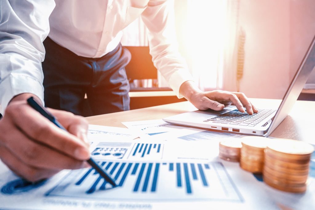 補助金申請の事業計画書を作成するため、資料やグラフを確認しながらノートパソコンを操作するビジネスパーソン