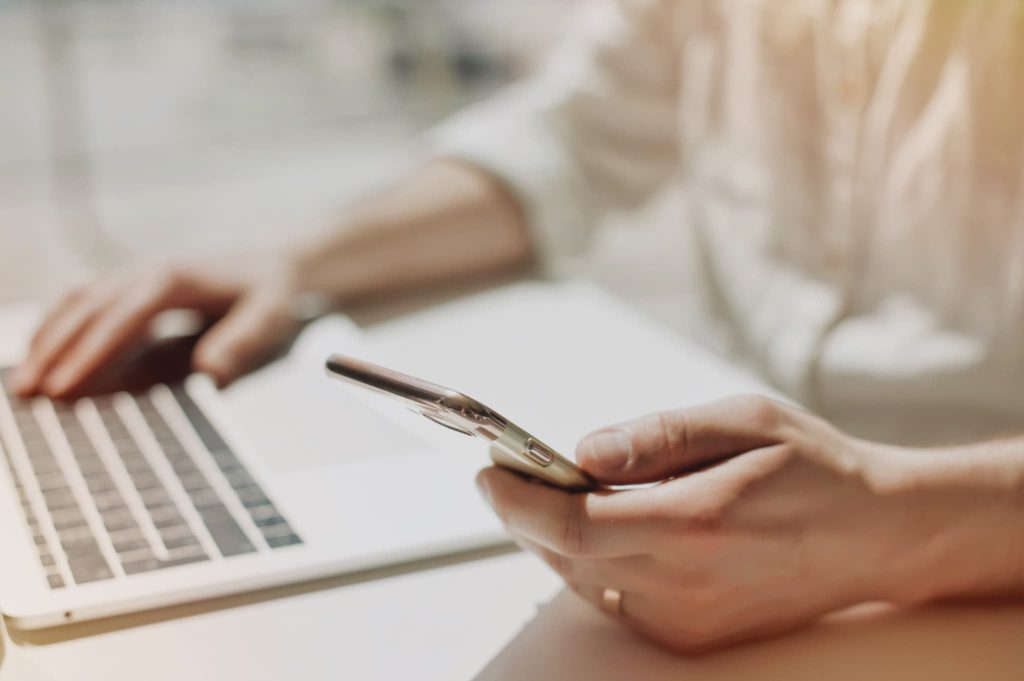 パソコンの前でスマートフォンを操作するビジネスパーソンの手元。経費削減に役立つアプリやクラウドサービスを活用して業務効率化を図る様子