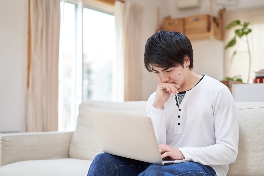 自宅のソファでノートパソコンを前に考え込む男性。ファクタリングの必要書類や審査の進捗を自宅から確認している様子