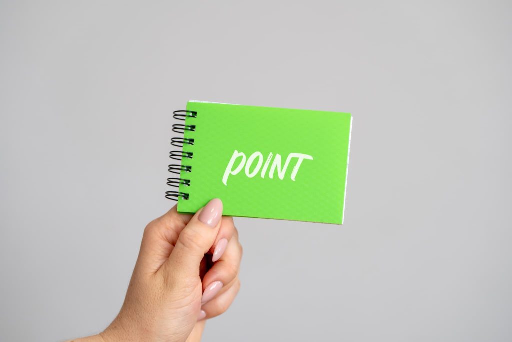 「POINT」と書かれた緑色の小さなノートを掲げる手。開業資金を抑えるための節約術や、助成金・補助金活用の重要ポイントを示すイメージ
