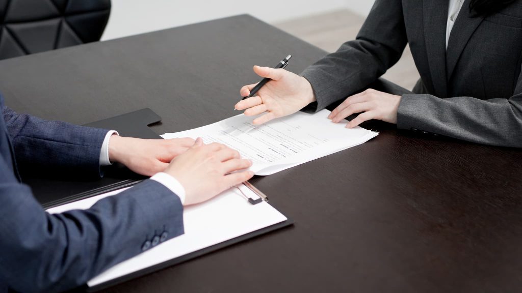 テーブルの上に広げられた契約書類と、説明を行うビジネスパーソンの手元。開業資金の融資実行や店舗・事務所の賃貸契約を交わす重要な手続きのイメージ