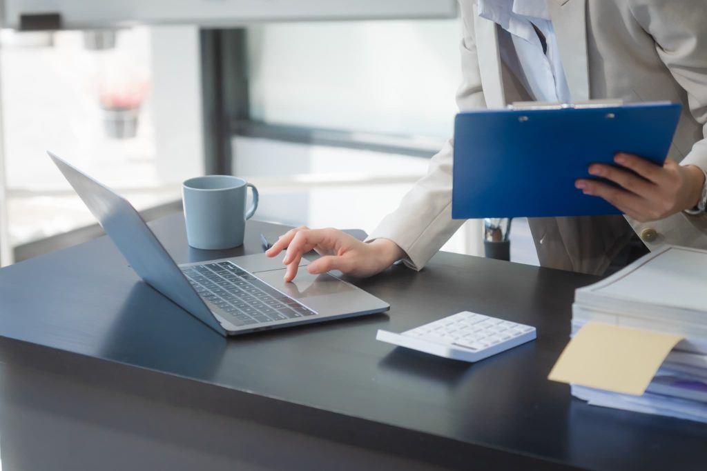 青いクリップボードを抱えながらノートパソコンで作業するオフィスワーカー。資金繰り表のテンプレートに入力し最新の財務状況を更新する事務作業