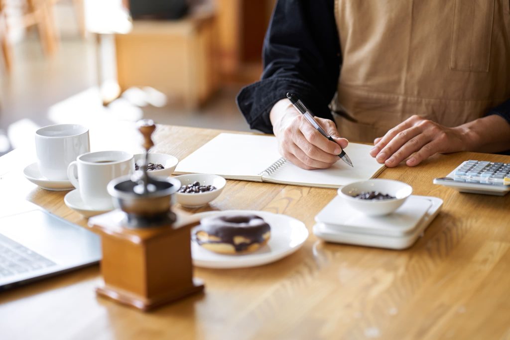 カフェの店主がノートに記録をつける手元。個人事業主が店舗運営の合間にファクタリングによる資金調達を検討し、収支を確認する様子