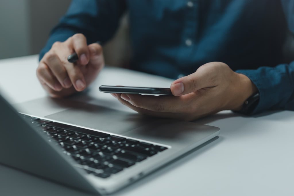 ノートパソコンの傍らでスマートフォンを操作するビジネスマン。資金繰りが苦しい際、早急に解決策を検索したり融資の申し込みをしたりする手元